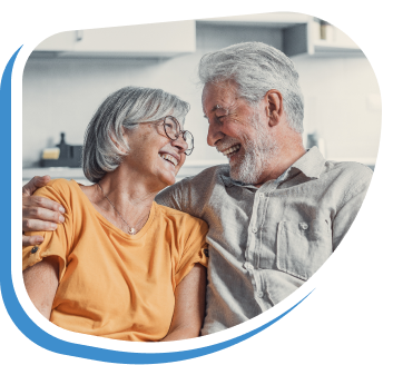 an older couple sits on a grey couch in their living room smiling and laughing to each other